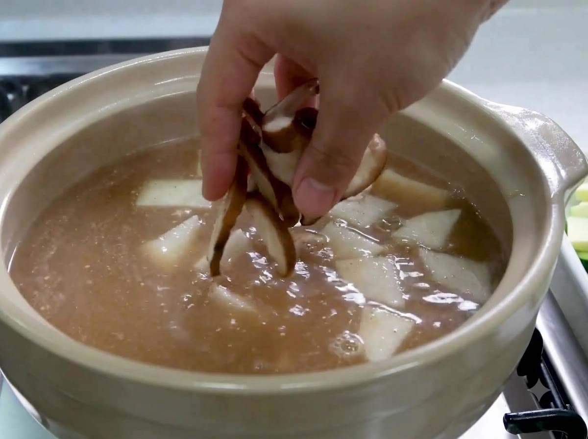 Una mano che fa cadere funghi shiitake affettati in una pentola contenente brodo e fette di ravanello.