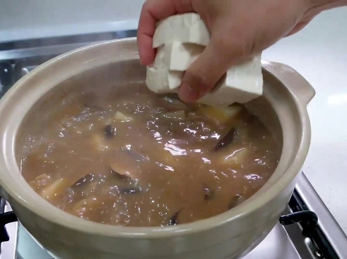 Una mano che fa cadere con attenzione cubetti di tofu bianco fresco in una pentola di stufato marrone bollente.