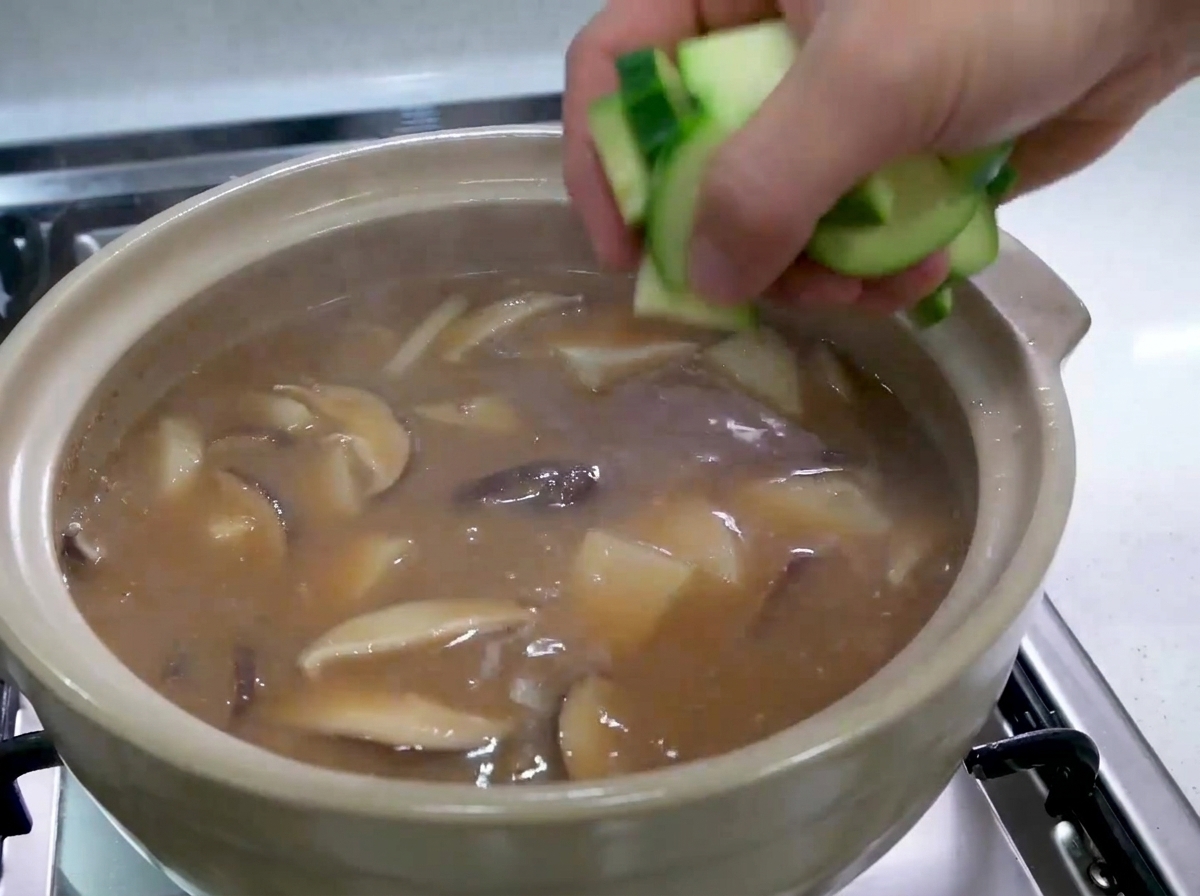Una mano che fa cadere pezzi tagliati di zucchine verdi crude in uno stufato coreano bollente.