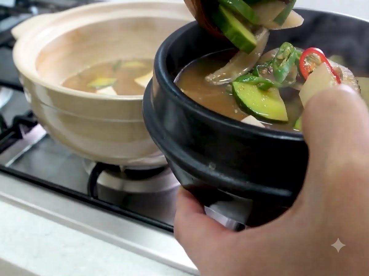 Un mestolo che trasferisce il Doenjang Jjigae caldo con zucchine e verdure in una ciotola da portata di pietra nera.
