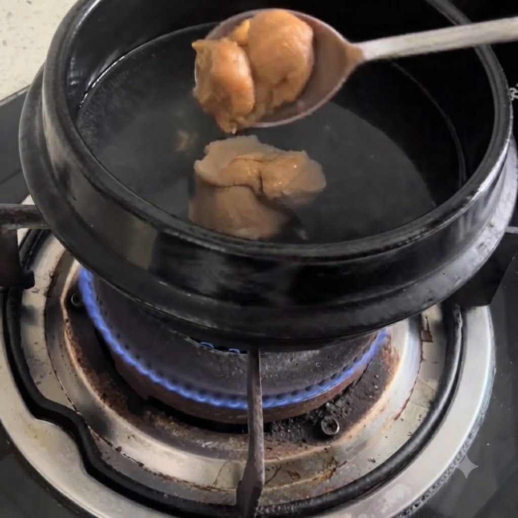 Un cucchiaio di pasta di soia che viene immerso in una pentola nera di acqua bollente su un fornello a gas.