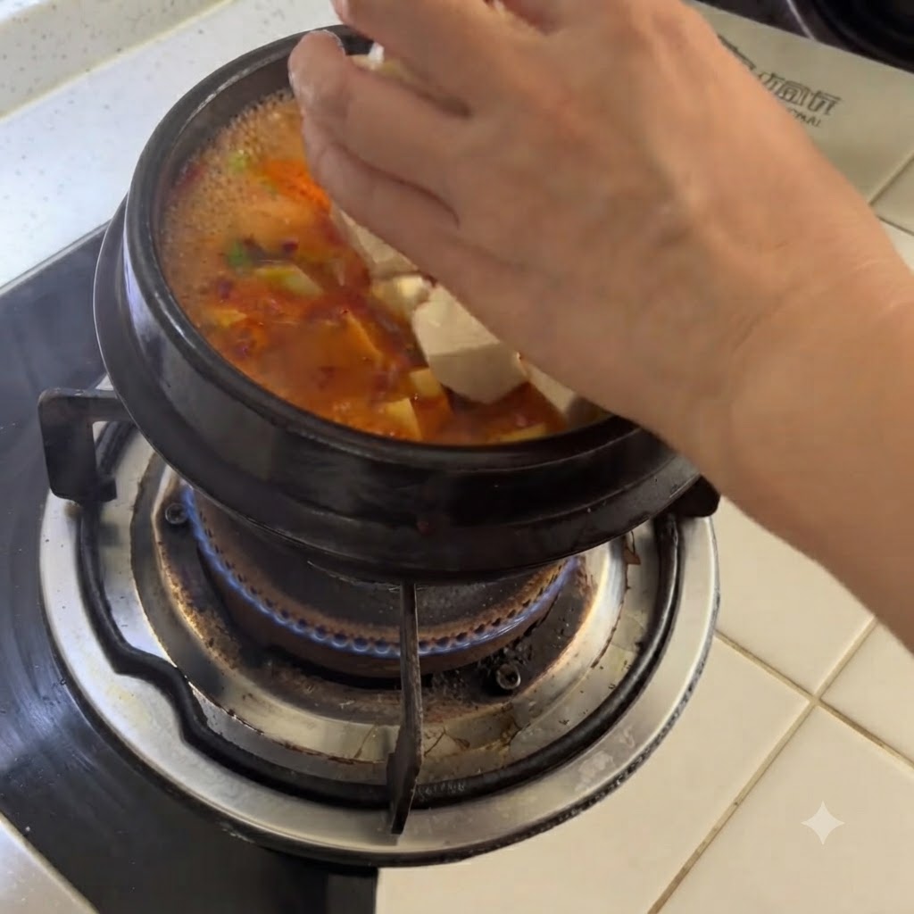 Tofu bianco a cubetti che viene fatto cadere a mano in una pentola di terracotta che sobbolle di zuppa piccante.