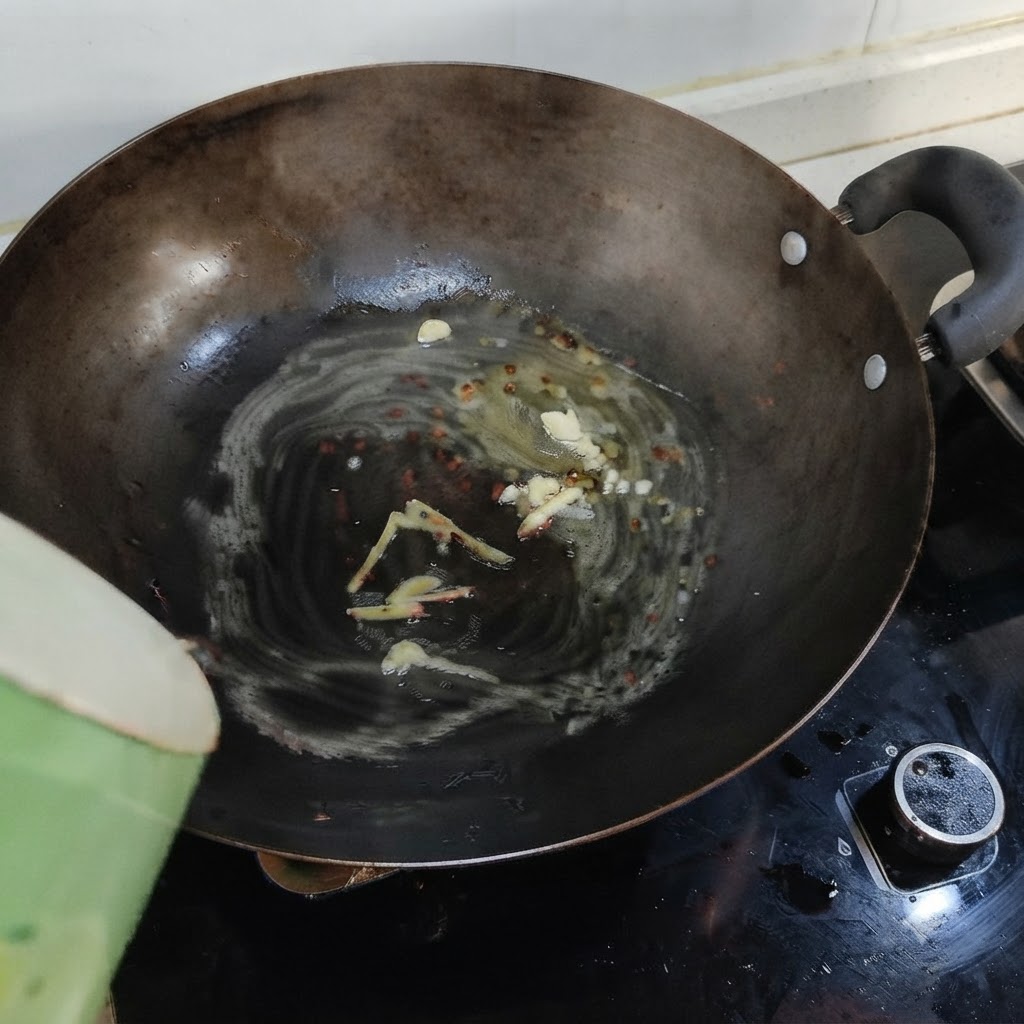 Acqua bollente versata da un contenitore verde in un wok contenente strutto sciolto, zenzero e aglio.