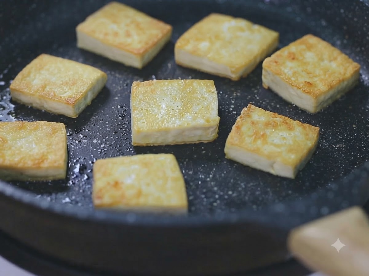 Diversi cubetti di tofu dorati che sfrigolano in una padella nera a fuoco basso.