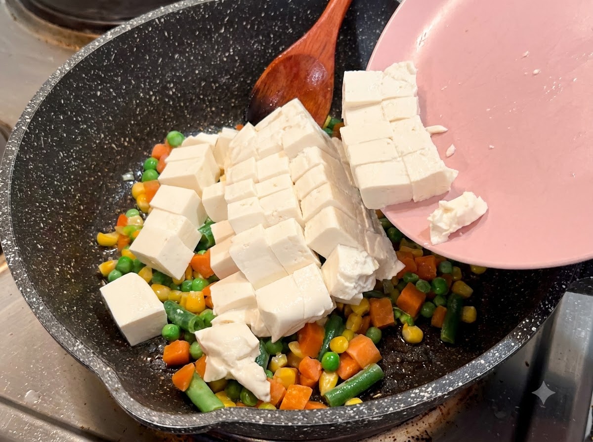 Tofu morbido a cubetti che scivola da un piatto rosa in una padella screziata con verdure miste.