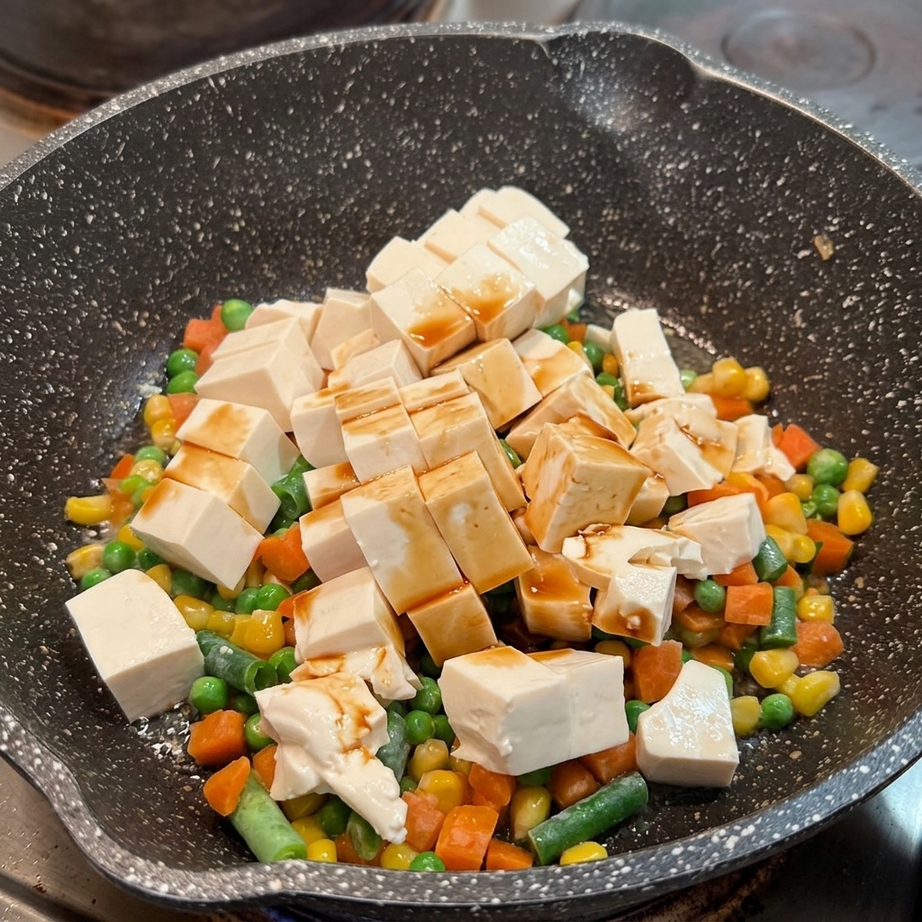 Salsa di soia distribuita sui cubetti di tofu morbido e verdure miste in una padella.
