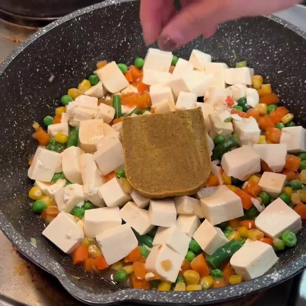 Una mano che posiziona un blocco di roux di curry marrone solido sul tofu e sulle verdure in una padella.