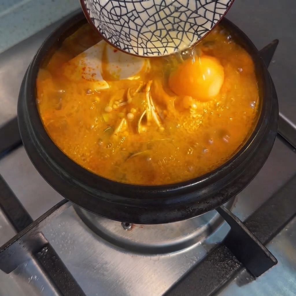 Un uovo crudo versato da una ciotolina nella zuppa di tofu coreana in ebollizione.