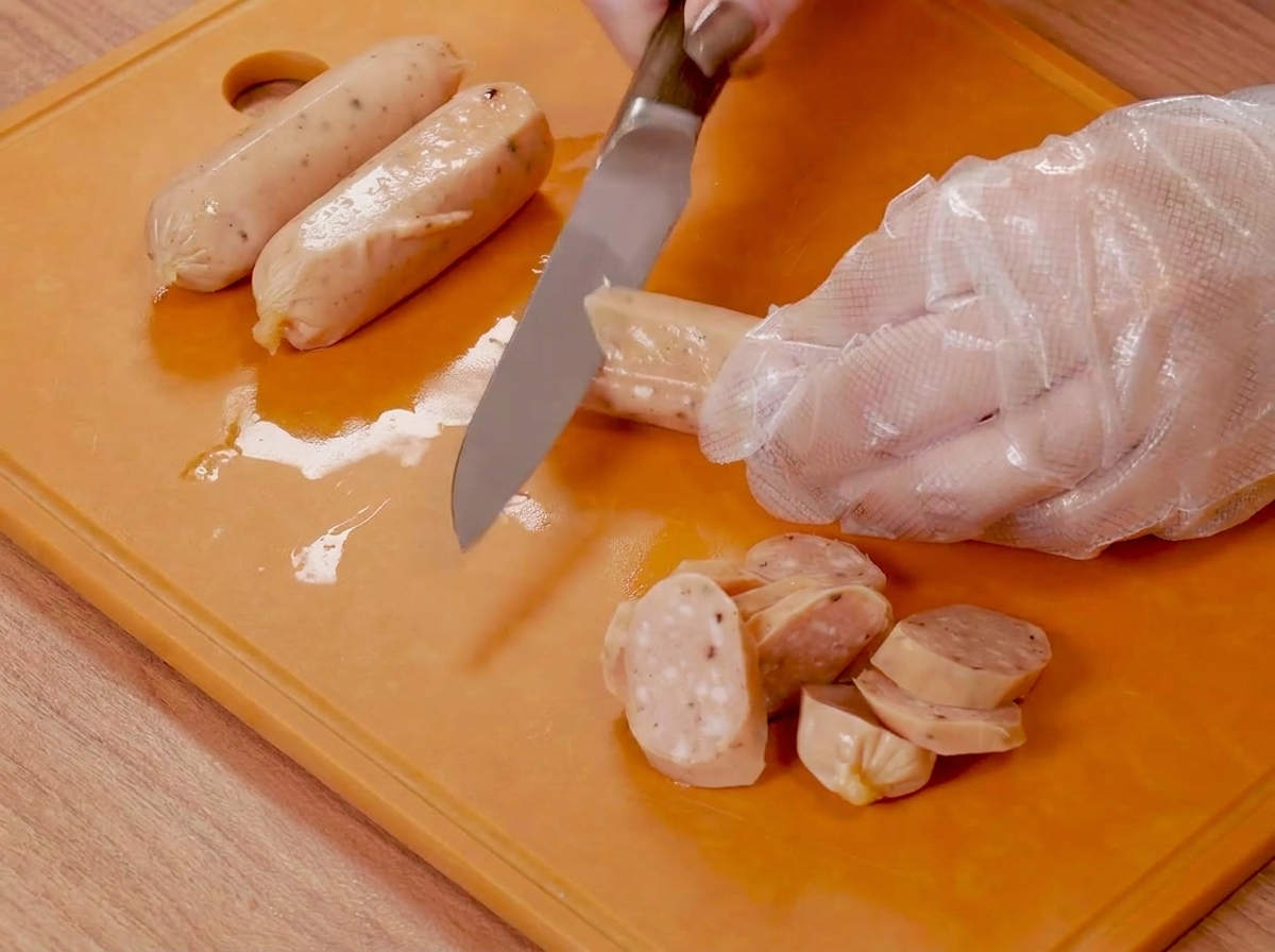 Mani con guanti di plastica che usano un coltello per incidere piccole salsicce su un tagliere arancione.