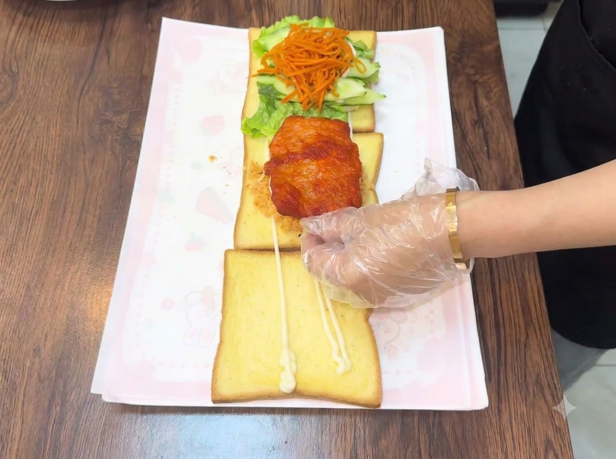 Una mano guantata che posiziona una cotoletta di pollo fritta calda sopra lassemblaggio di pane a strati.