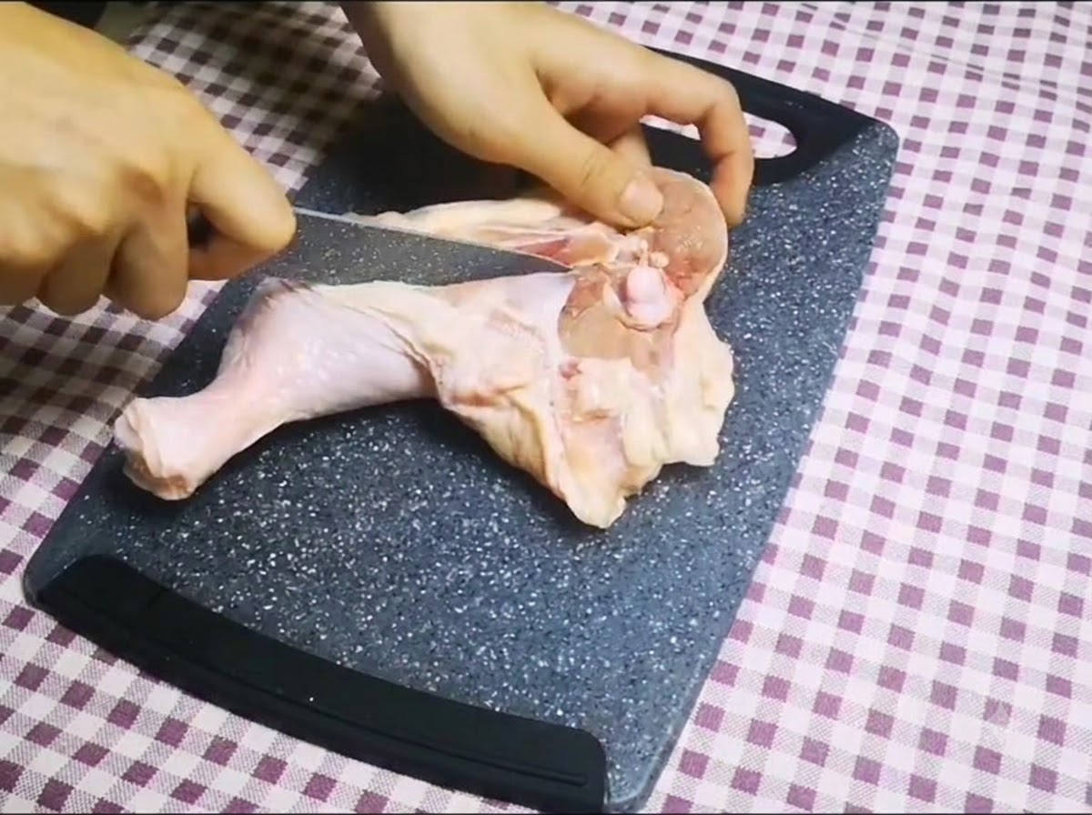 Un coltello affilato che taglia lungo losso di una coscia di pollo cruda su un tagliere per rimuovere losso.