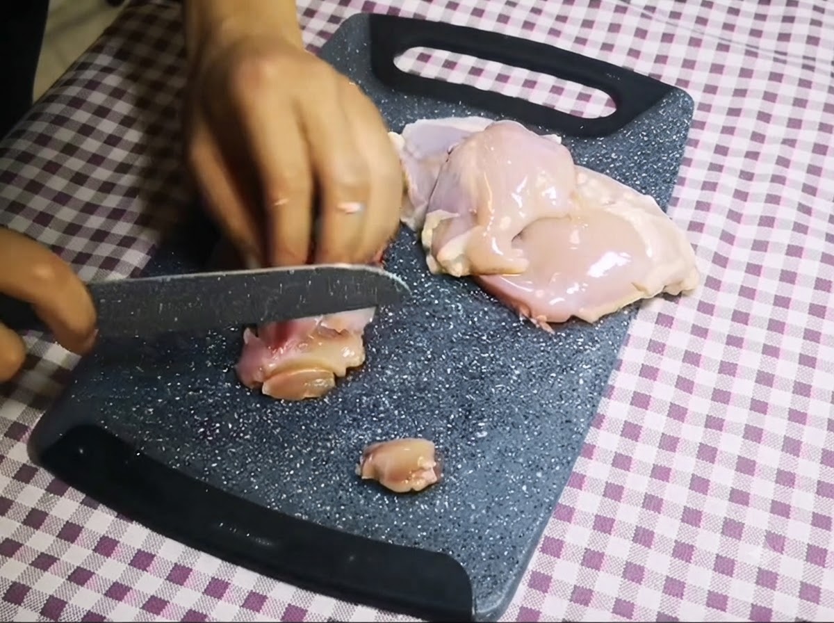 Un coltello che taglia pollo crudo disossato in piccoli pezzi su un tagliere grigio.