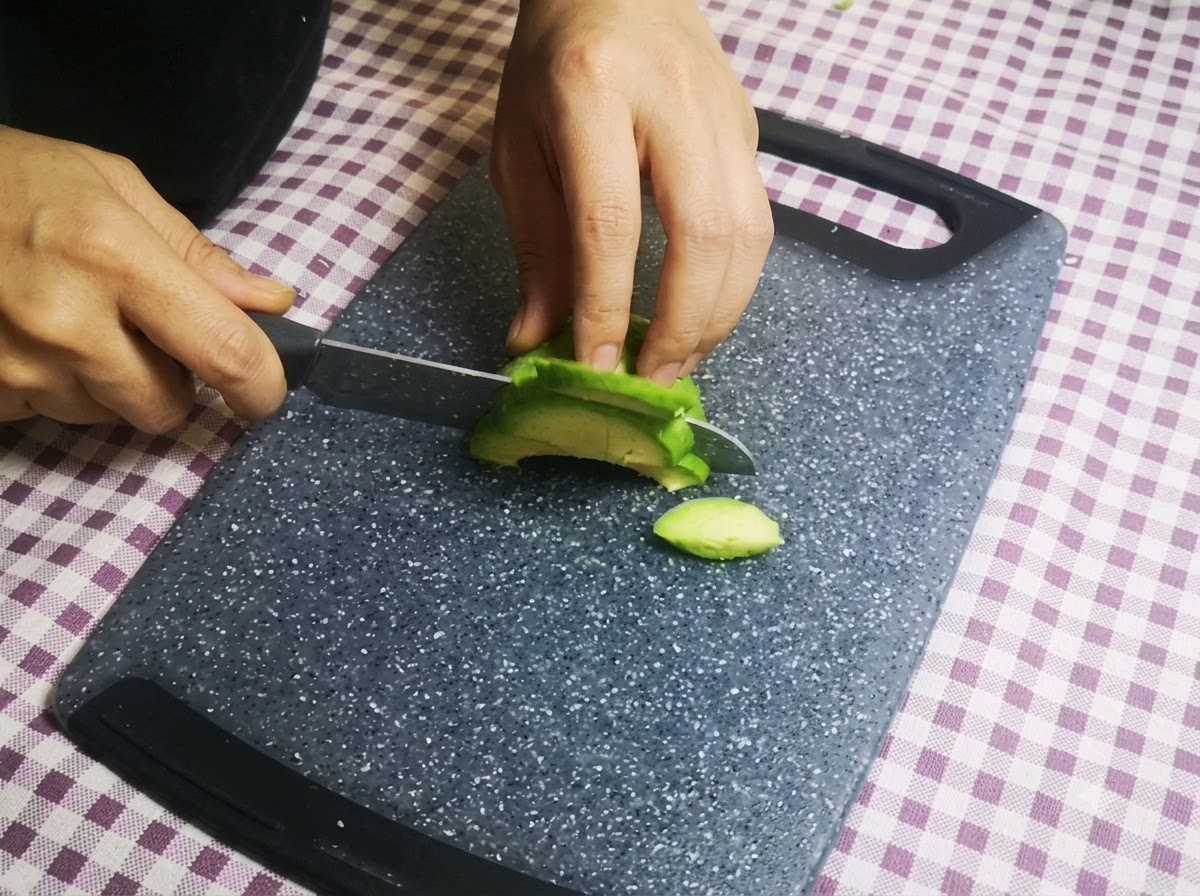 Mani che tagliano una metà di avocado verde in fette sottili con un coltello su un tagliere.
