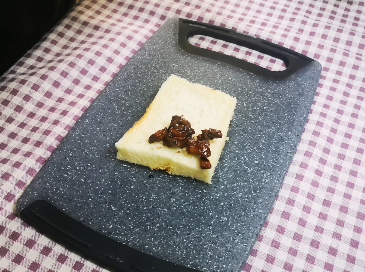 Primo piano di pezzi di pollo glassati scuri disposti ordinatamente su una singola fetta di pane bianco in cassetta poggiata su un tagliere grigio.
