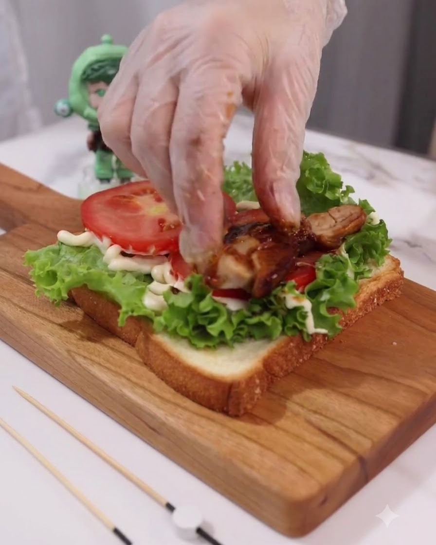 Mano con guanto che posiziona fette di pollo teriyaki cotto sugli strati di pomodoro e lattuga.