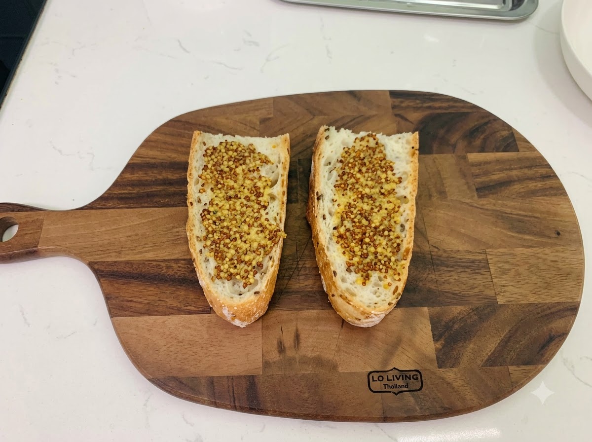 Due fette di pane rustico su un tagliere di legno, entrambe spalmate uniformemente con senape in grani.
