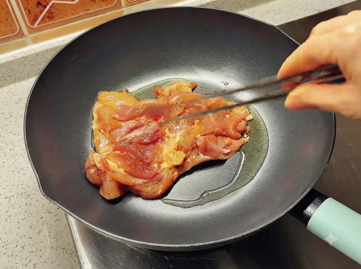 Un singolo pezzo di coscia di pollo marinato che viene inserito in una padella calda unta dolio usando delle pinze.
