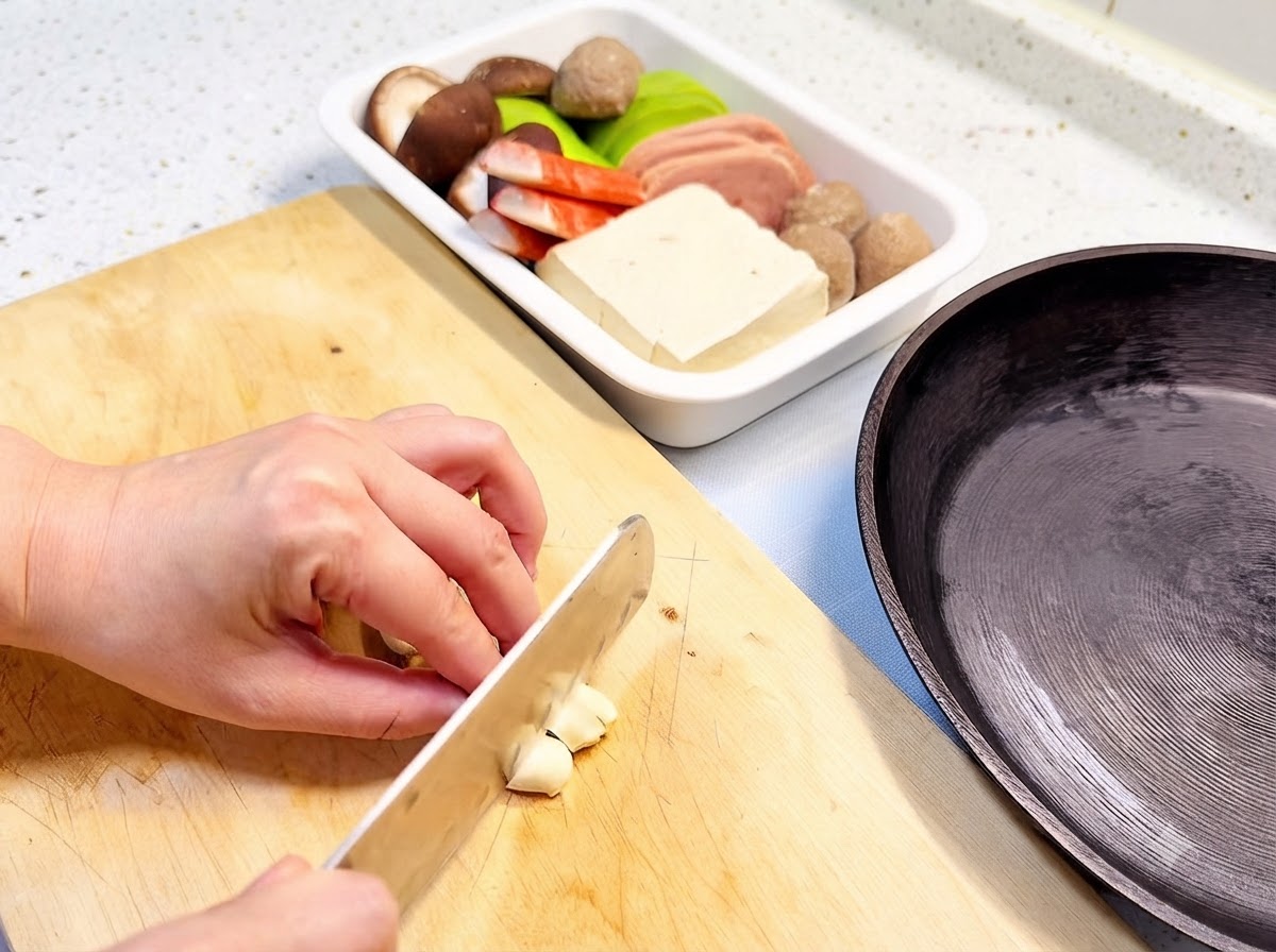 Spicchi daglio affettati su un tagliere di legno accanto a una padella e a un vassoio di ingredienti.