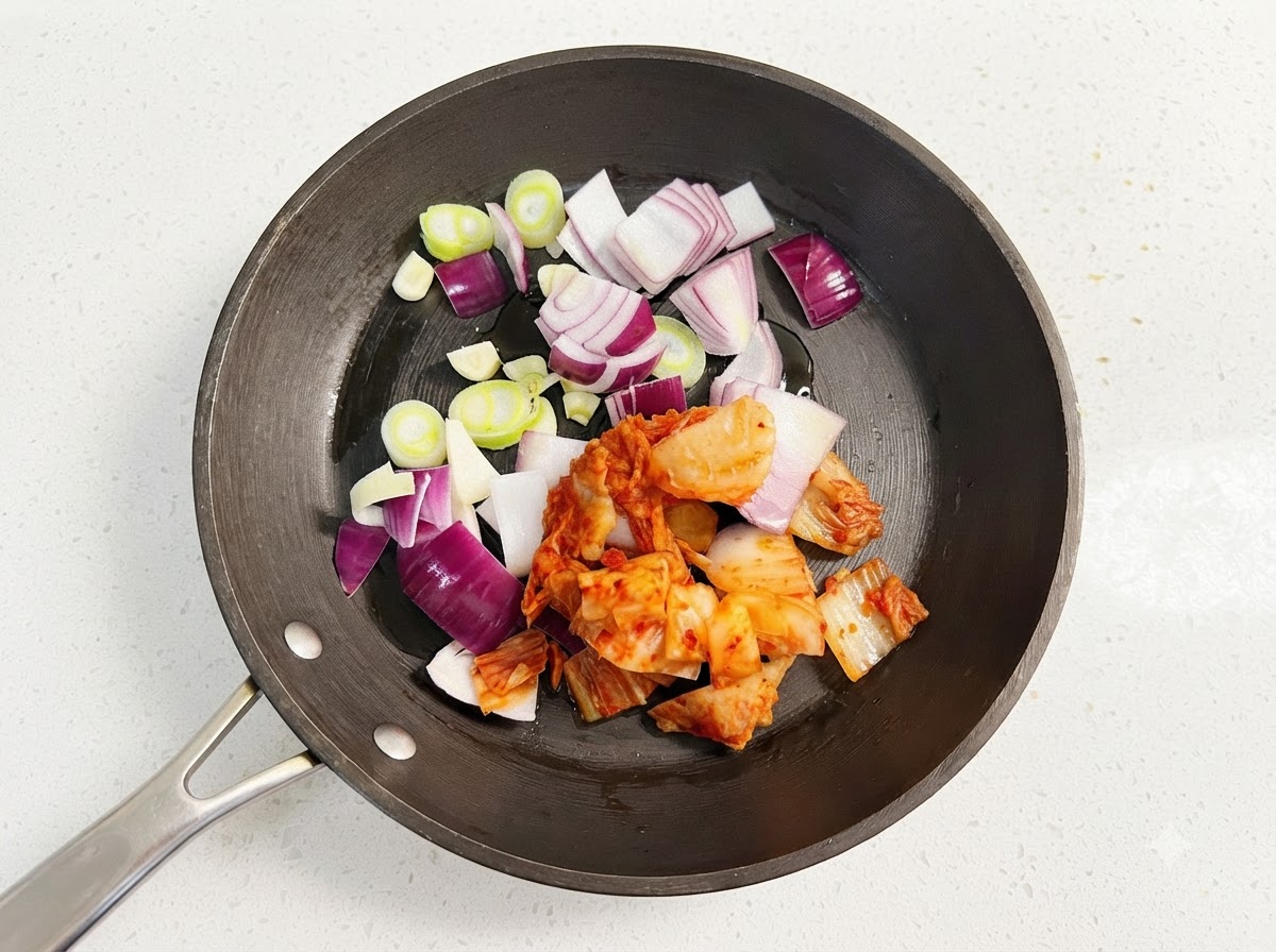 Fette di cipolla viola, rondelle di scalogno verde e kimchi rosso tritato che sfrigolano in una padella nera con olio.