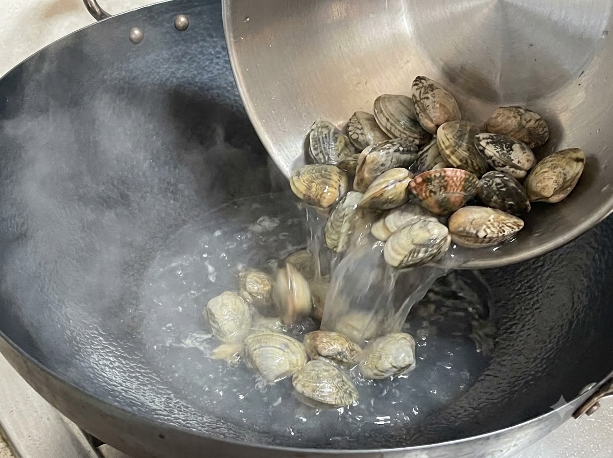 Vongole fresche versate da una ciotola di metallo in un wok fumante.