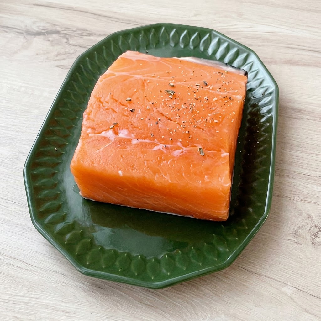Un trancio spesso di salmone fresco su un piatto verde, spolverato generosamente con sale marino grosso e pepe nero macinato.