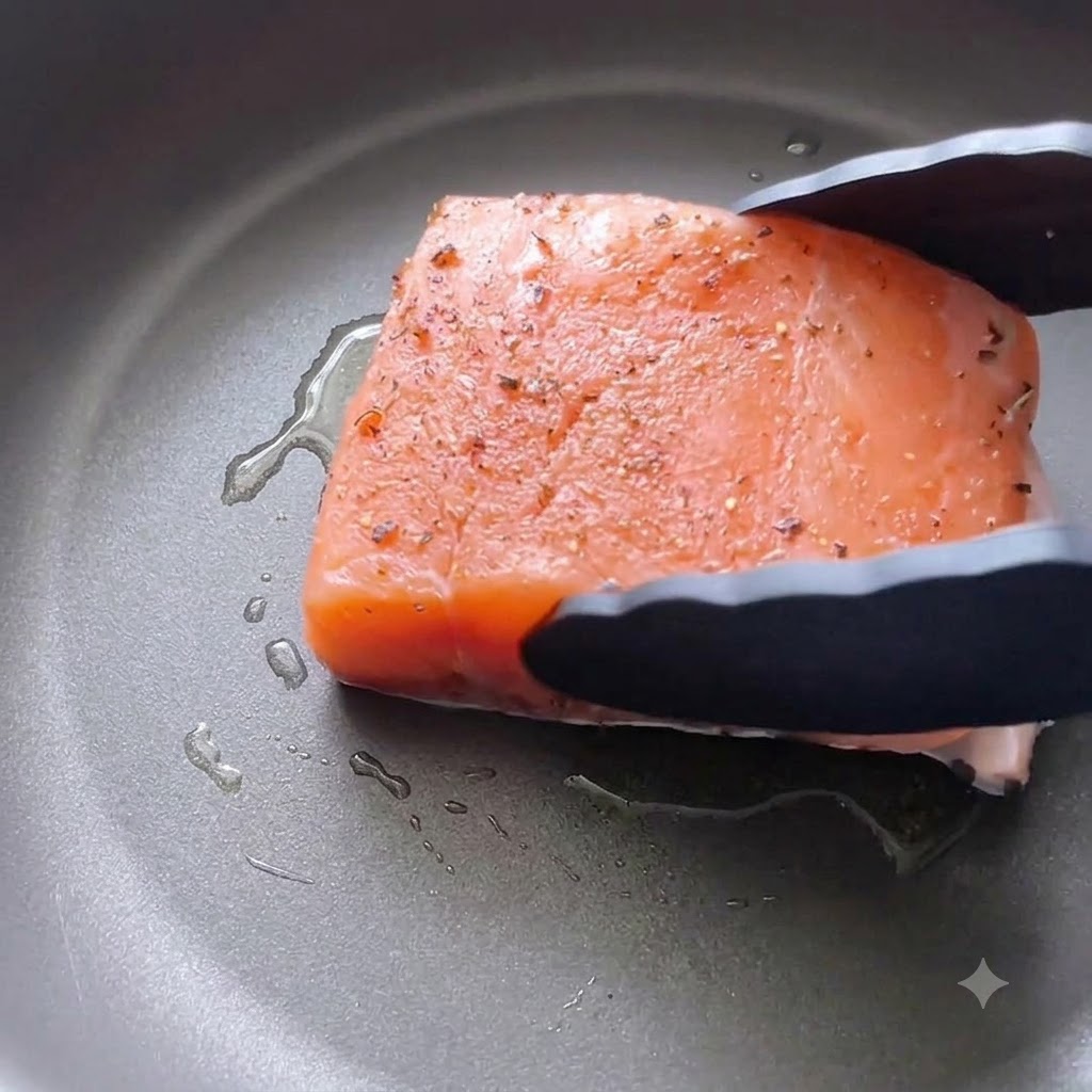 Un filetto di salmone condito viene adagiato con la pelle rivolta verso il basso in una padella oliata usando delle pinze da cucina nere.