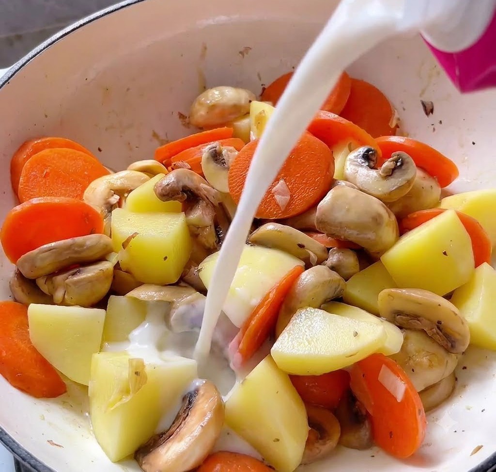 Liquido versato da un cartone rosa in una pentola piena di cubetti di patate, fette di carota e funghi.