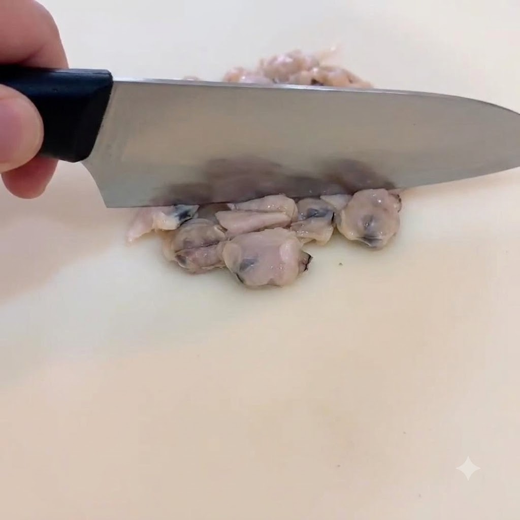 Primo piano di una mano che trita delicatamente al coltello le vongole cotte su un tagliere bianco.