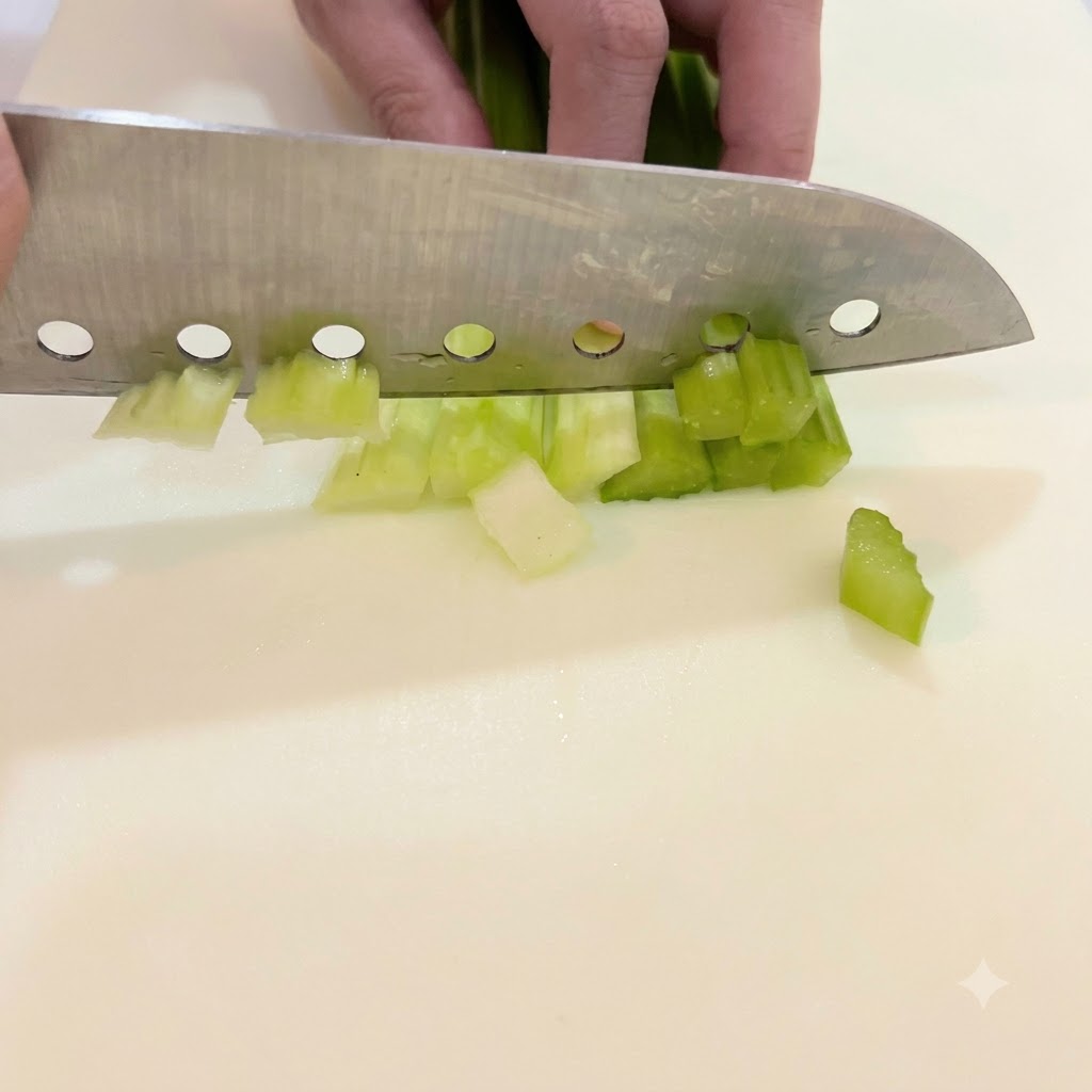 Mano che guida un grande coltello per tagliare coste di sedano verde brillante a pezzetti su un tagliere.