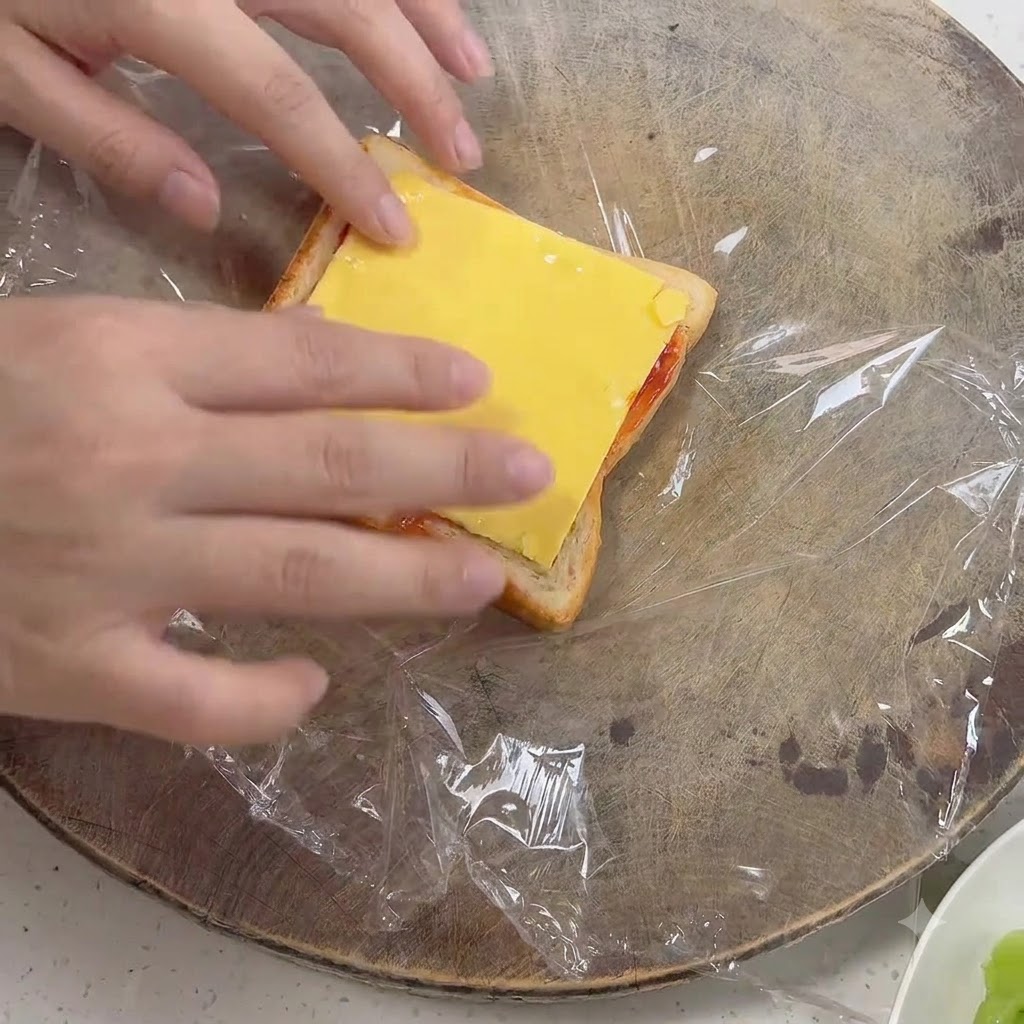 Mani che posizionano una fetta quadrata di formaggio giallo su un pezzo di toast coperto di salsa.