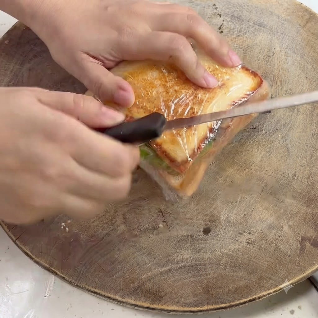 Un coltello da cucina che taglia diagonalmente attraverso un panino avvolto in plastica su un tagliere di legno.