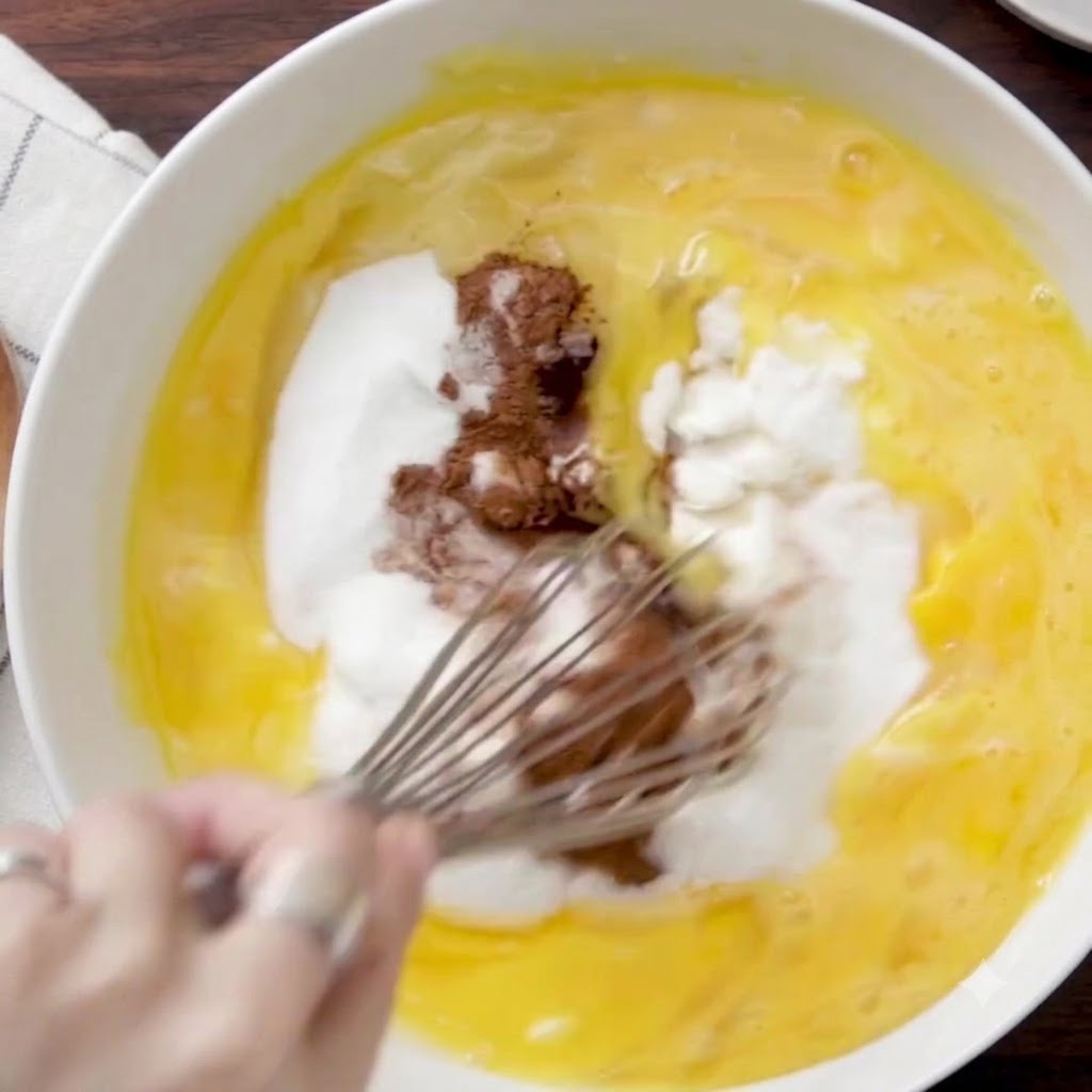 Una frusta a filo che mescola ingredienti umidi, zucchero e cannella allinterno di una grande ciotola bianca.