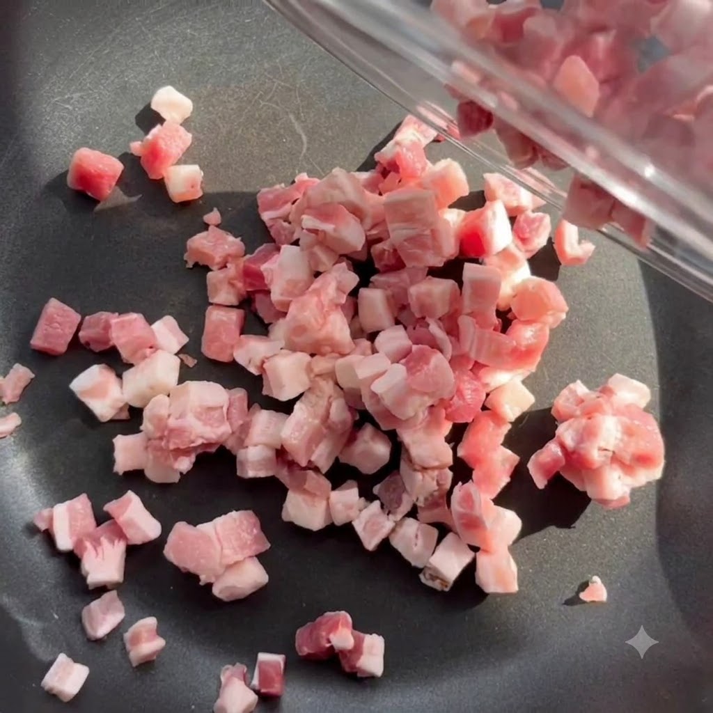 Pezzi di pancetta a cubetti che cuociono e sciolgono il grasso in una padella nera a fuoco medio.