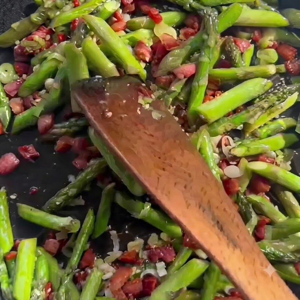 Brodo liquido chiaro versato da un contenitore in una padella sfrigolante piena di pezzi di asparagi verdi affettati e pancetta.