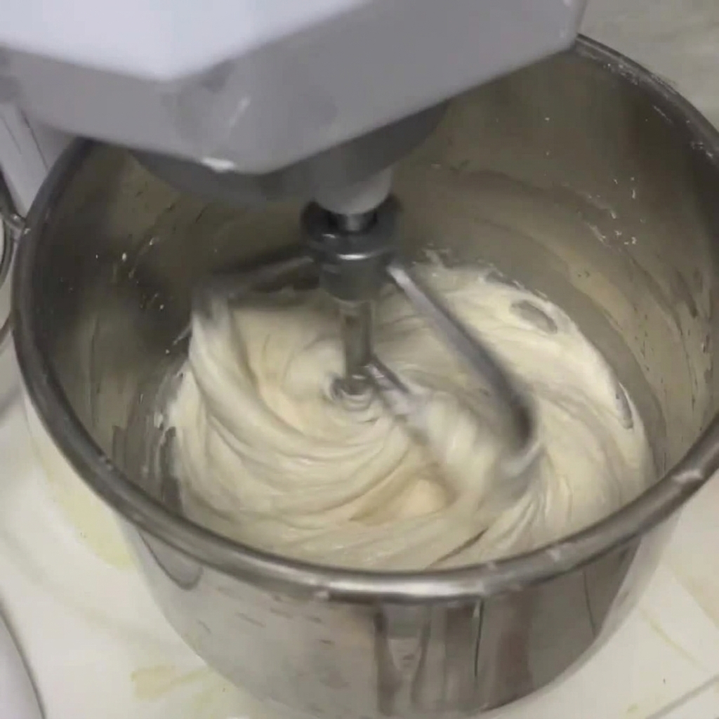 Impasto bianco che si unisce al liquido allinterno della ciotola di una grande planetaria in acciaio inox usando il gancio impastatore.