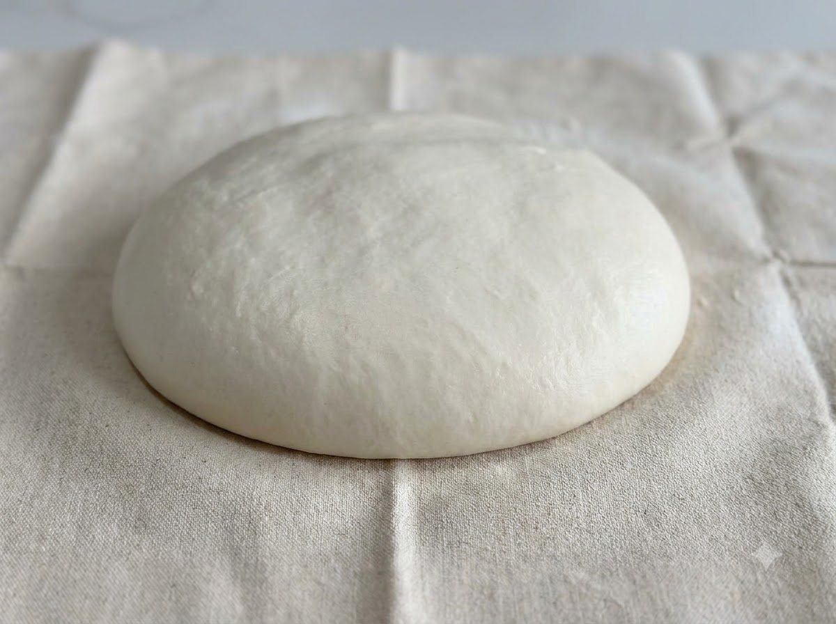 Una cupola liscia e arrotondata di impasto bianco adagiata su una superficie di stoffa bianca infarinata.