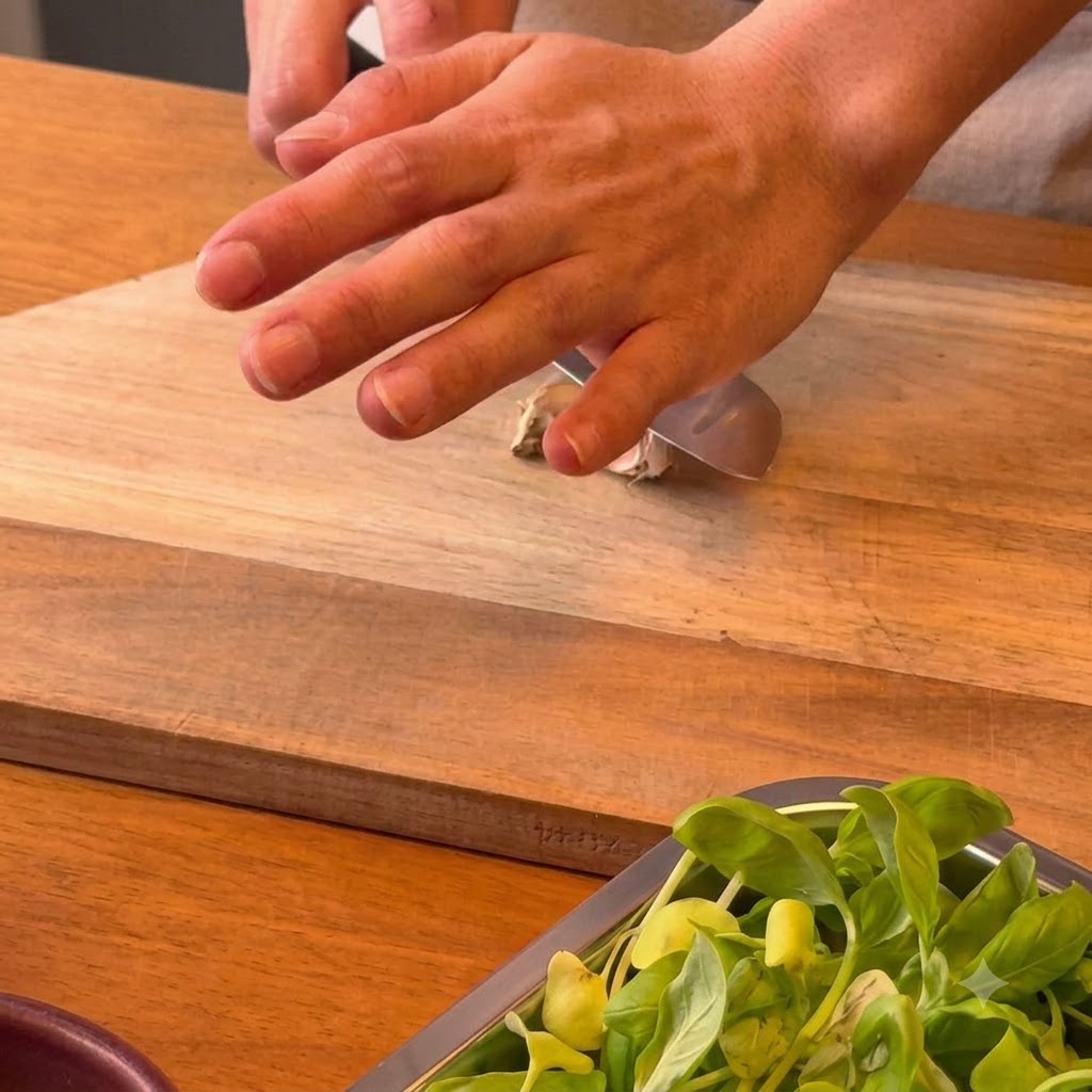 Una mano che preme il lato piatto di un coltello da chef contro uno spicchio daglio su un tagliere di legno.