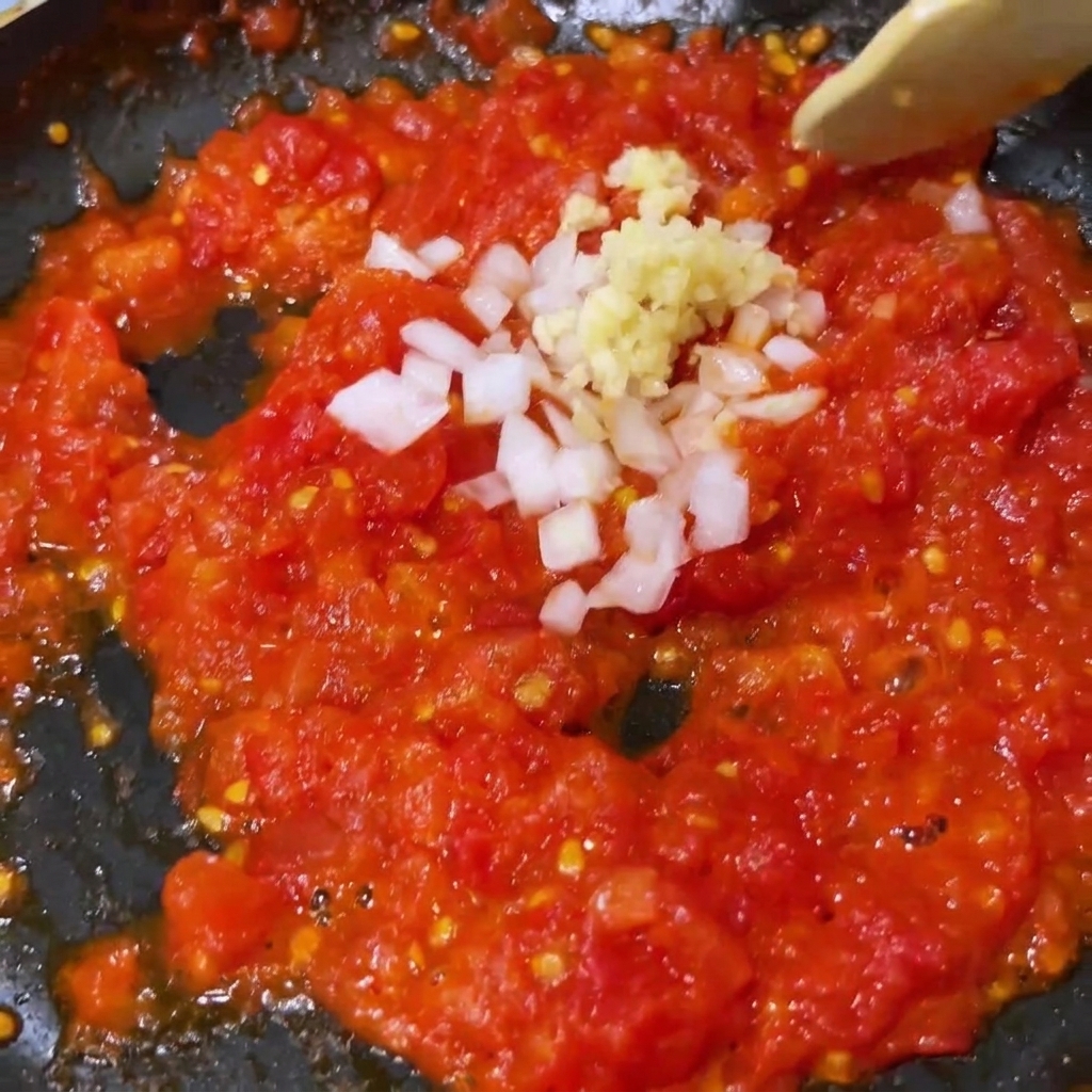 Aglio tritato e cipolla bianca mescolati ai pomodori schiacciati e ammorbiditi in padella.