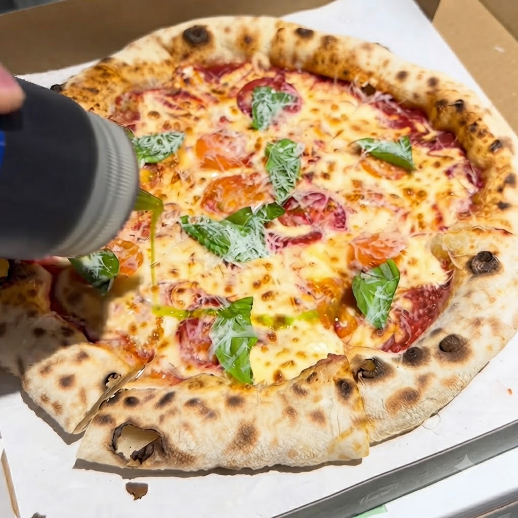 Una bottiglia che versa un filo di olio extravergine doliva su una pizza appena sfornata condita con basilico e formaggio in una scatola.
