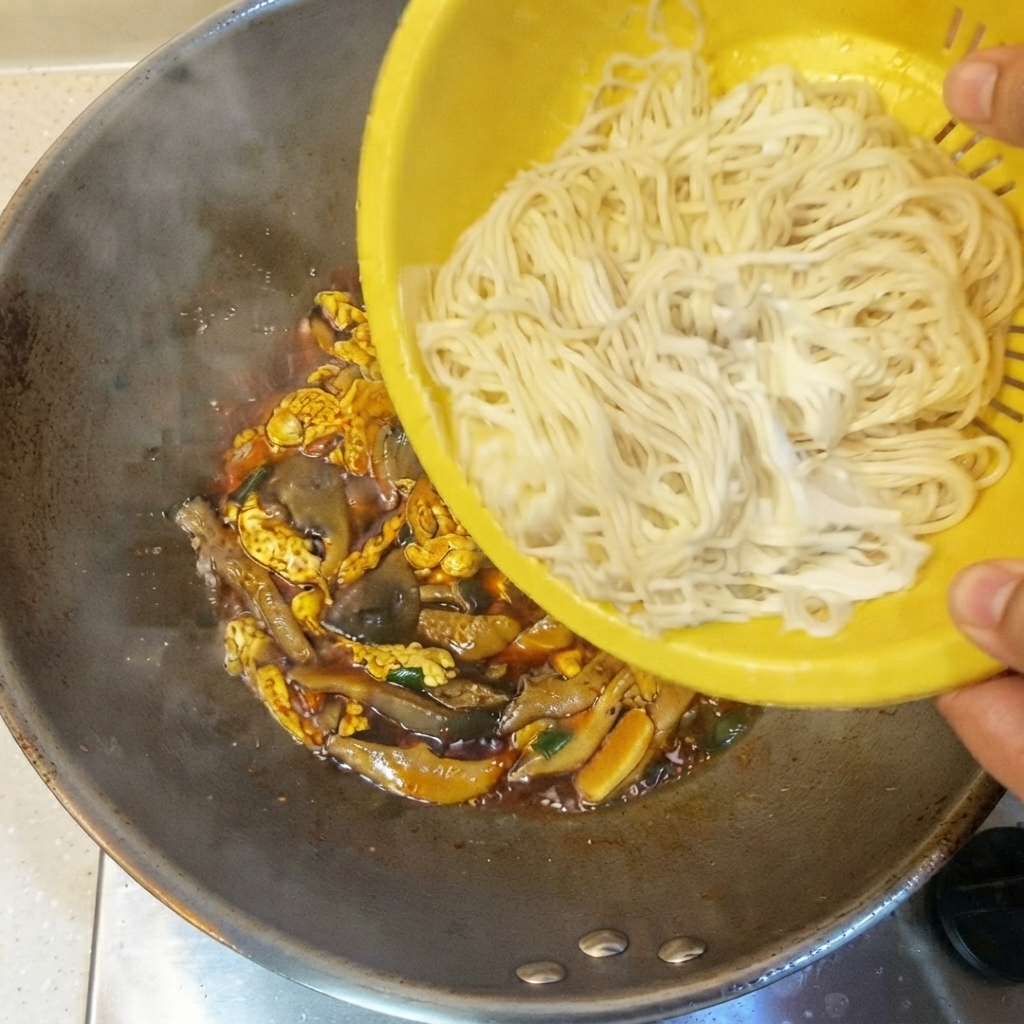 Noodles sottili cotti versati da un colino giallo nel wok contenente la salsa e gli ingredienti.