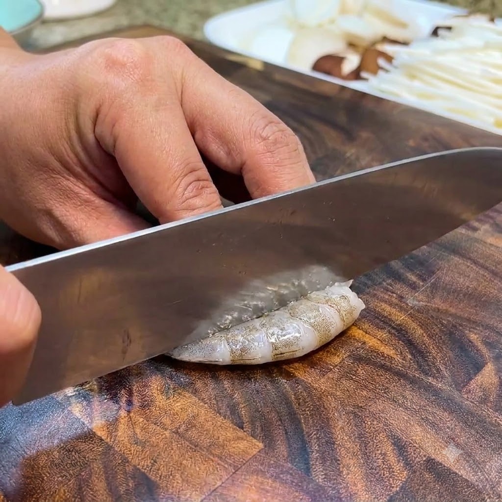 Gamberi crudi tagliati e puliti con un coltello su un tagliere di legno.