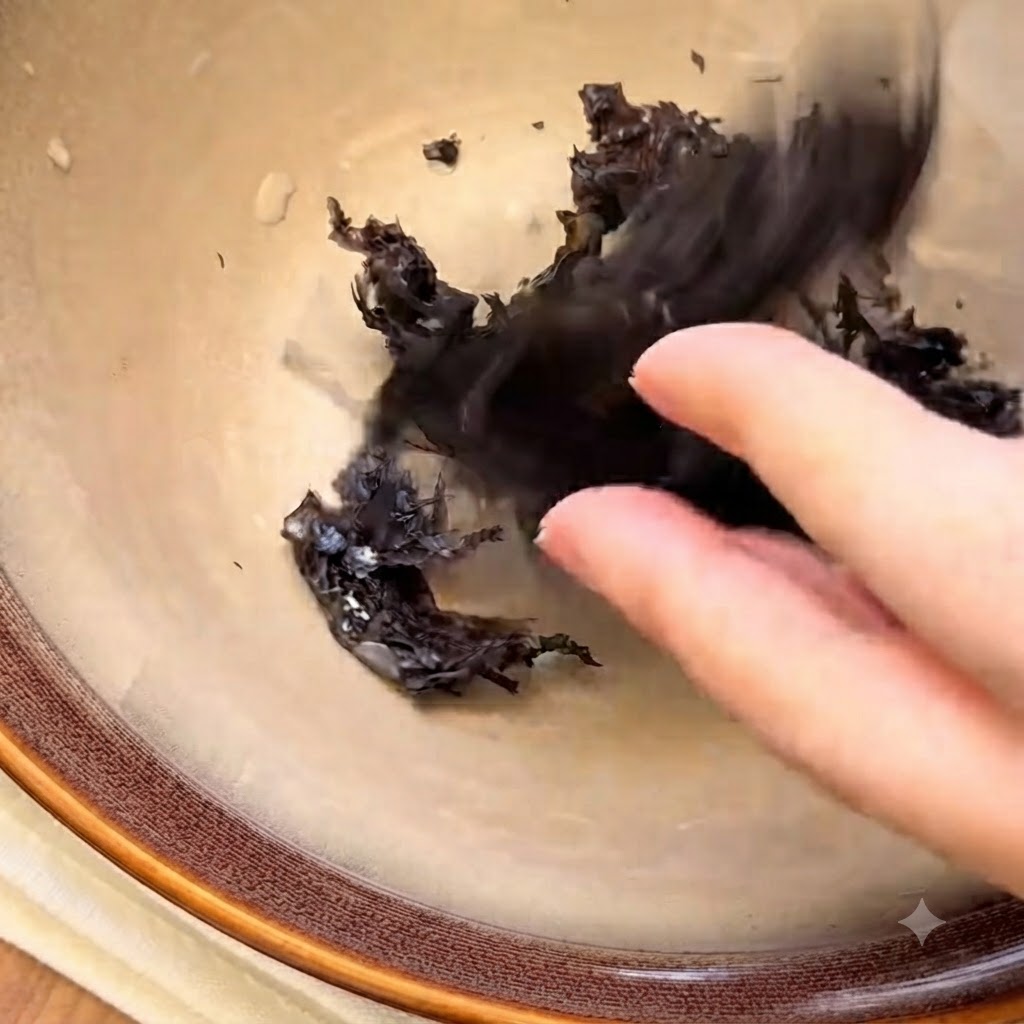 Pezzi di alga disidratata e semi di sesamo bianco disposti sul fondo di una ciotola di ceramica vuota.