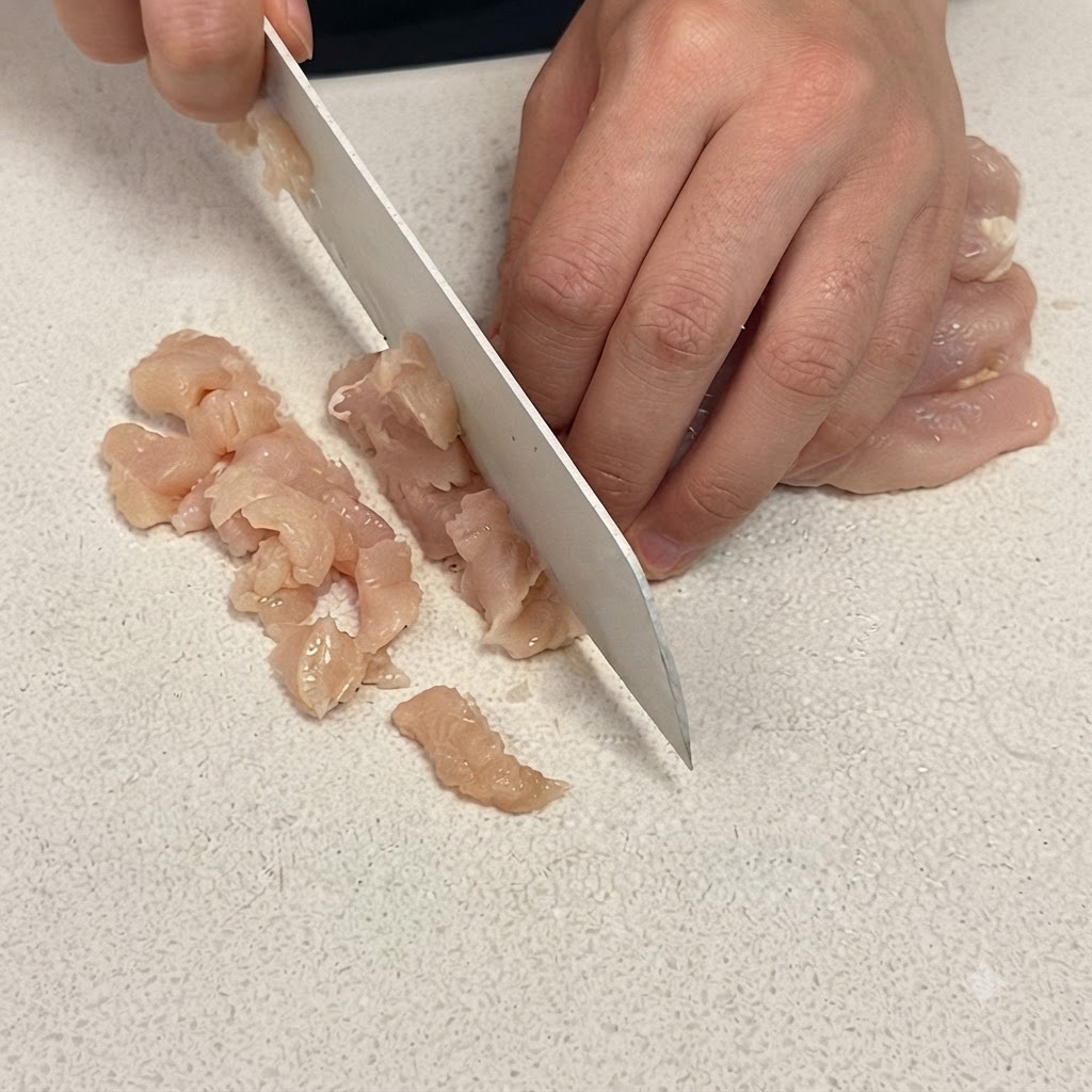 Un primo piano di un petto di pollo tagliato a cubetti piccoli su un tagliere bianco con un affilato coltello da chef.