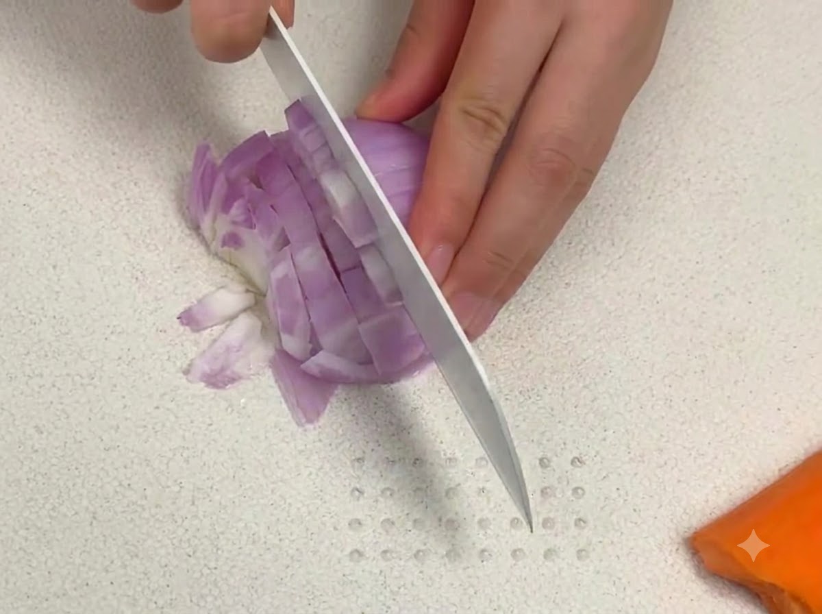 Un coltello affilato che taglia cipolla rossa a dadini su un tagliere bianco vicino a un pezzo di carota.