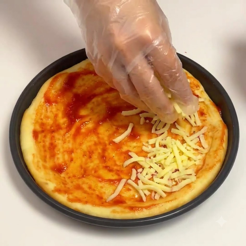 Una mano guantata che sparge mozzarella grattugiata sulla base di salsa di pomodoro in una teglia per pizza rotonda.