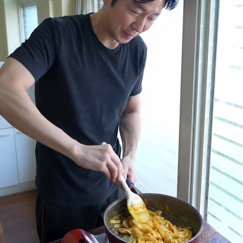 Una persona che mescola i noodle larghi in una padella con una spatola bianca mentre regola la consistenza della salsa.