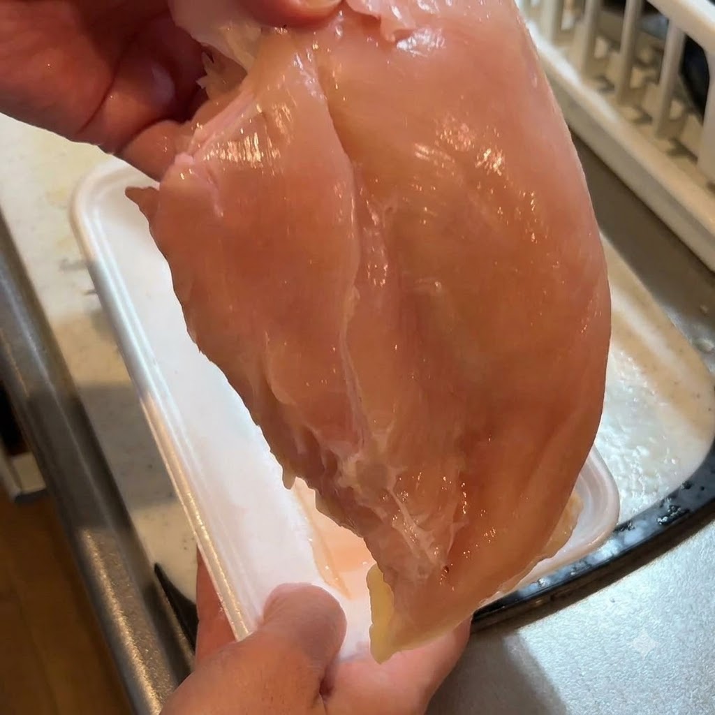 Un petto di pollo crudo tenuto in alto mentre la pelle viene rimossa con un coltello.