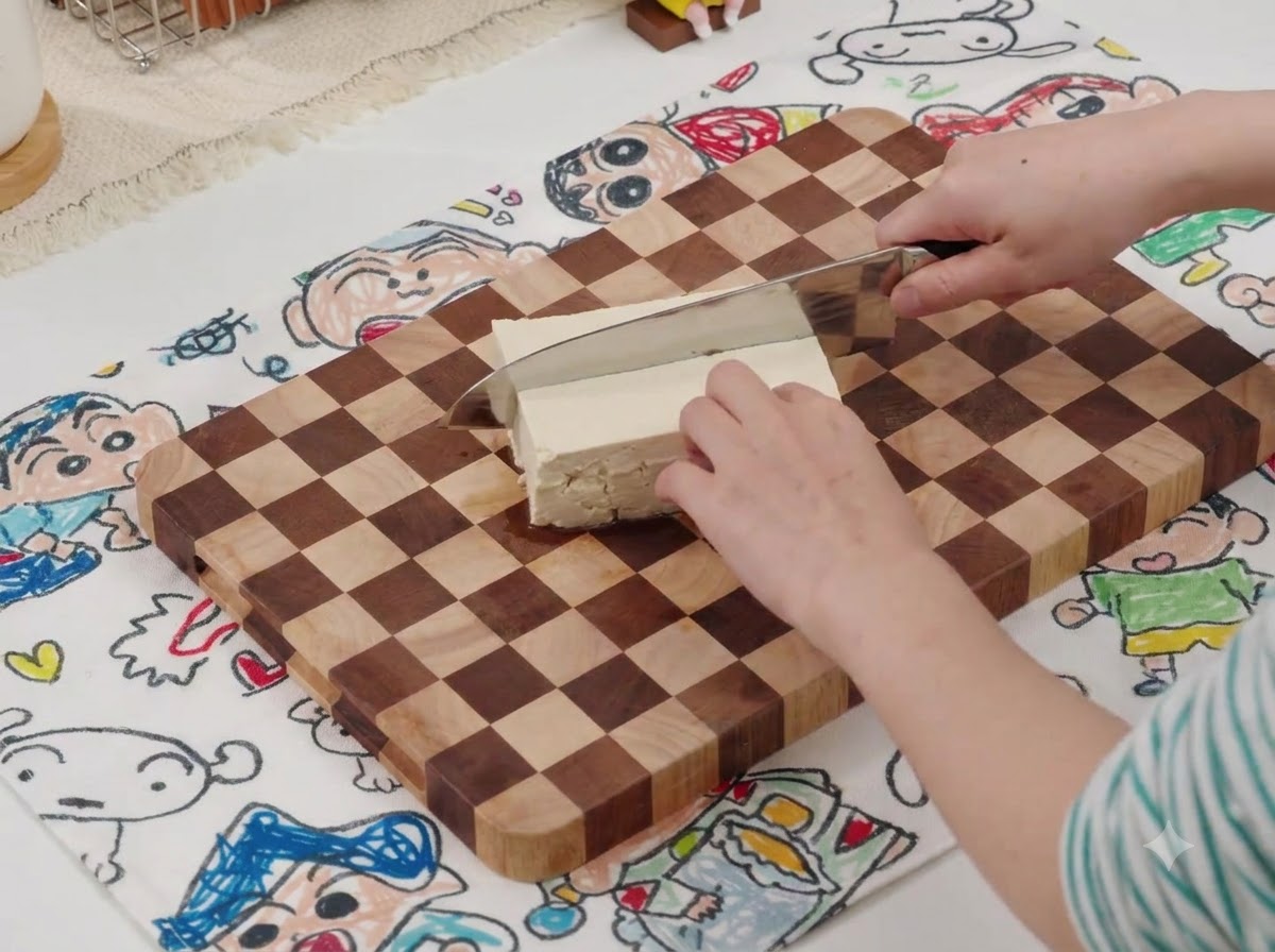 Un coltello che taglia un blocco di tofu bianco su un tagliere di legno con un tappetino fantasia sotto.