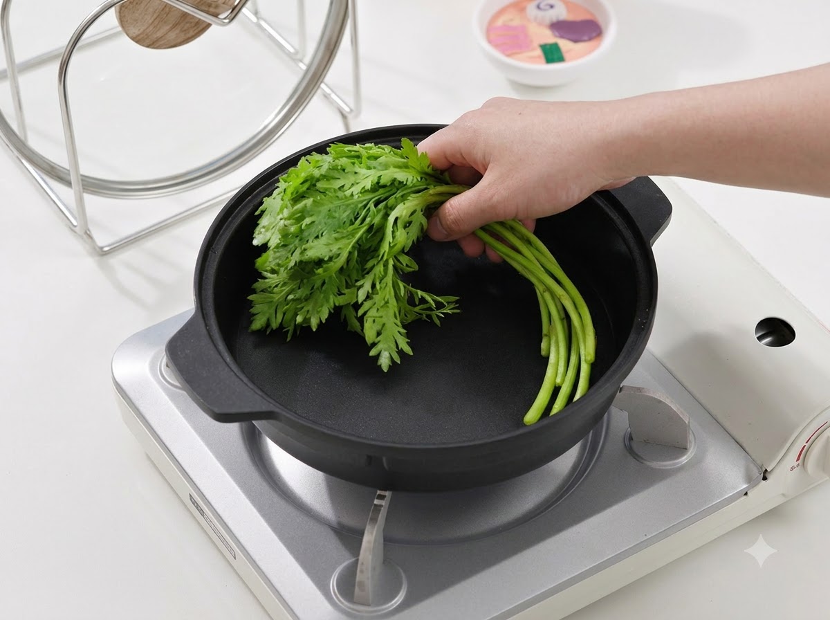 Una mano che posiziona un mazzetto di verdure a foglia verde fresca in una padella nera su un fornello portatile.