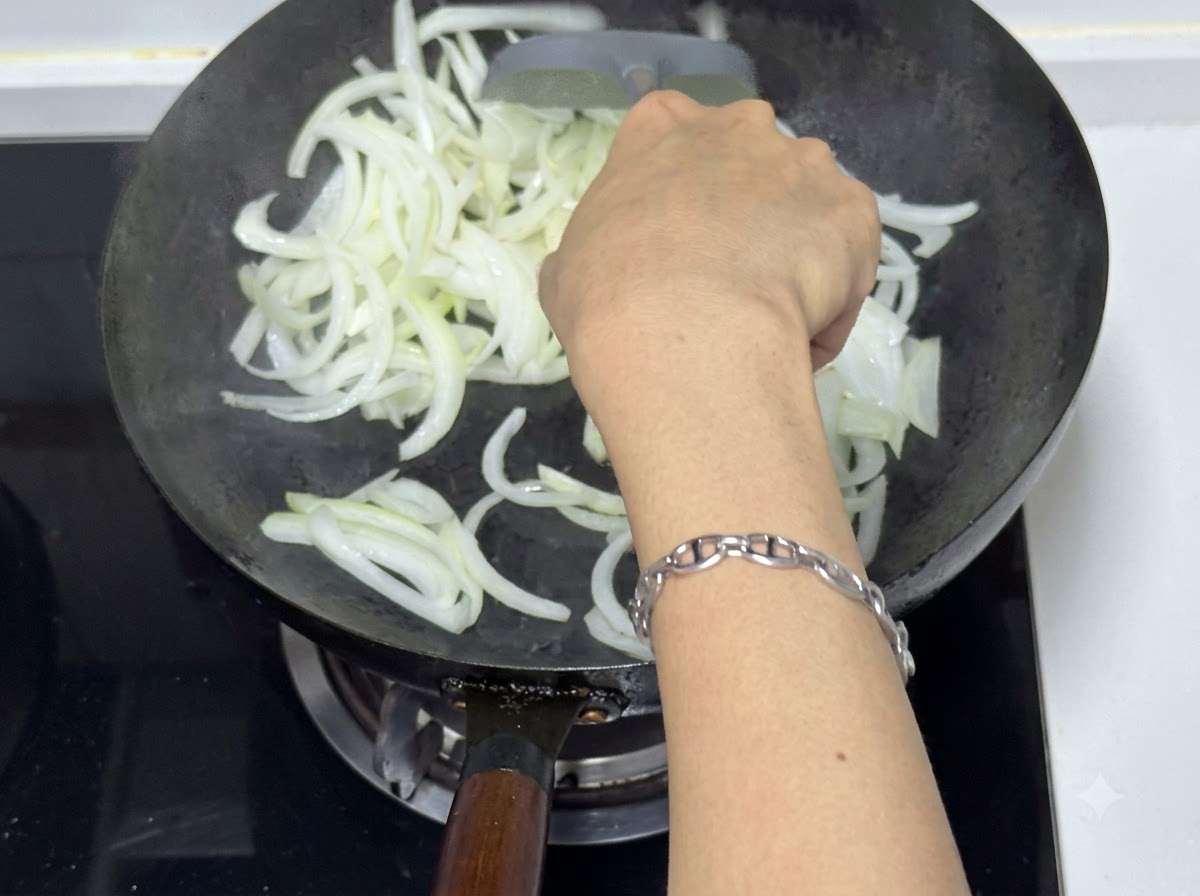 Una persona che usa una spatola per saltare le cipolle affettate in un wok, mostrando pezzi traslucidi e leggermente dorati.